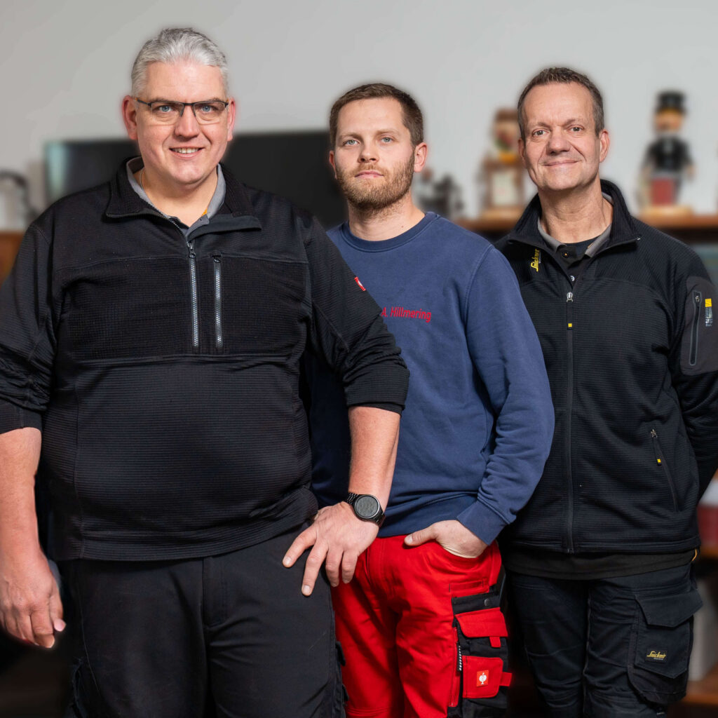 Drei Mitarbeiter von Clausen Haustechnik stehen gemeinsam für ein Gruppenfoto beisammen, frontal zur Kamera, in Arbeitskleidung und mit freundlichem Ausdruck.