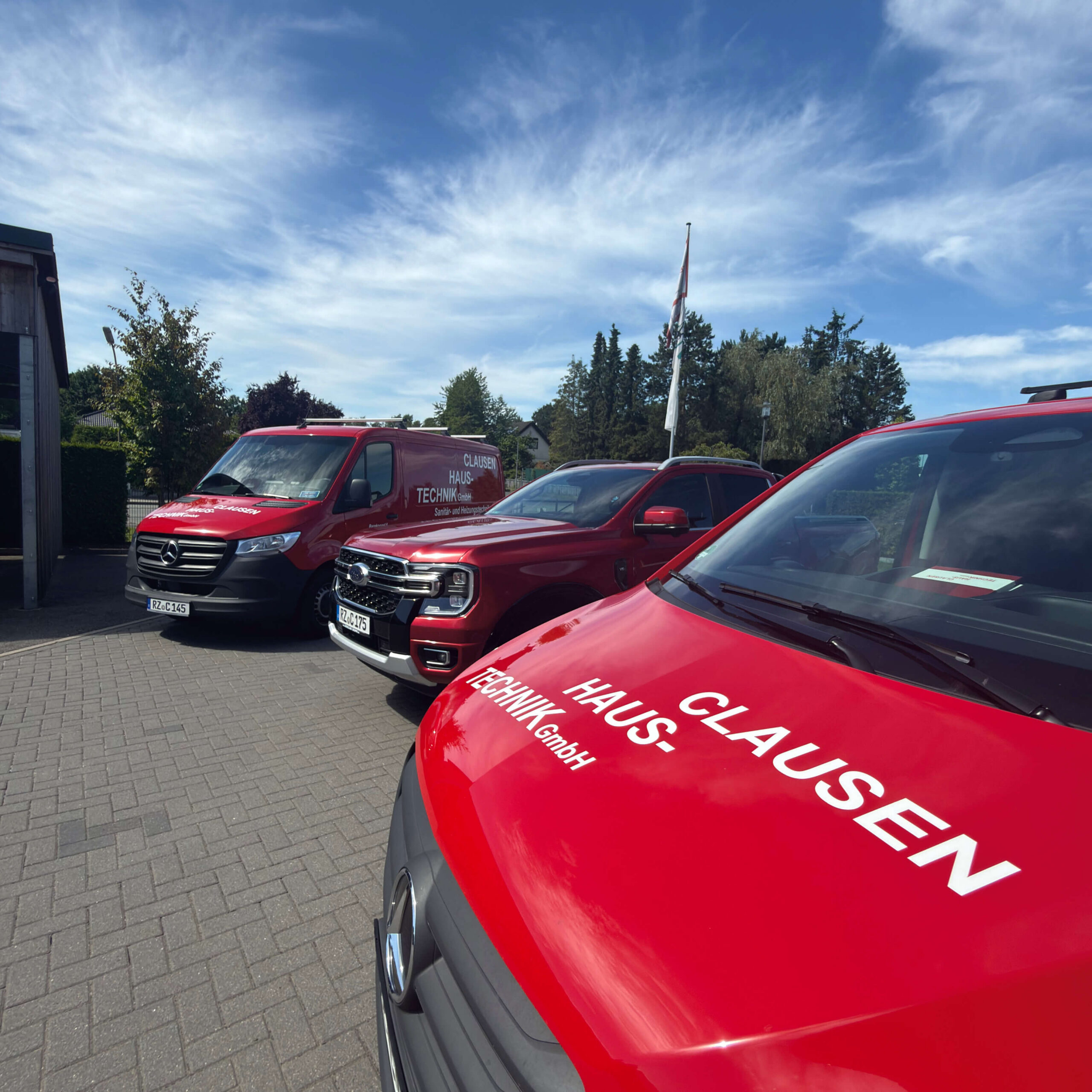 Drei rote Einsatzfahrzeuge von Clausen Haustechnik stehen auf dem Firmengelände. Im Vordergrund ist ein Transporter mit Firmenlogo zu sehen, dahinter ein Ford Ranger und ein Mercedes Sprinter unter blauem Himmel.